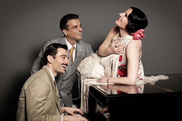 Una mujer vestida de coplera ríe recostada sobre un piano. Dos hombres la miran sonrientes. Uno de ellos toca el piano.